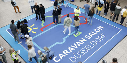 Aufnahme von oben: blauer Bodenaufkleber mit Schrift 'CARAVAN SALON D&Uuml;SSELDORF', stilisierte Wohnwagen-Grafik und mehrere Personen in einer Halle
