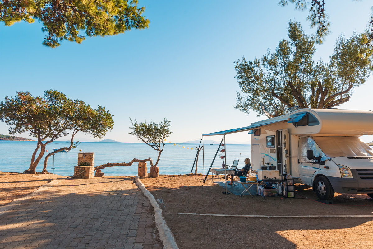 Wohnmobil mit ausgefahrener Markise, Campingtisch und St&uuml;hlen unter B&auml;umen am Meer; gepflasterter Weg und Meer im Hintergrund