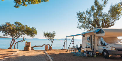 Wohnmobil mit ausgefahrener Markise, Campingtisch und St&uuml;hlen unter B&auml;umen am Meer; gepflasterter Weg und Meer im Hintergrund