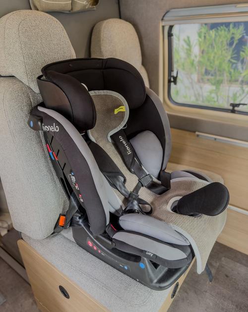 Schwarz-grauer Kindersitz mit Fünf-Punkt-Gurt auf hellem Fahrzeugsitz, Fenster und Holzfläche im Hintergrund