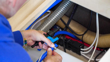 Hände einer Person halten ein blaues Kabel in einem offenen Schrankfach mit flexiblem Lüftungsschlauch, roten und weißen Leitungen und schwarzen Kabelkanälen