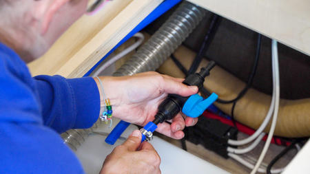 Zwei Hände in blauem Ärmel halten unter einer Arbeitsplatte ein schwarzes Kunststoffteil mit blauem Hebel, neben weißen und roten Schläuchen, einer silbernen Flexleitung und einer Metall‑Schlauchschelle
