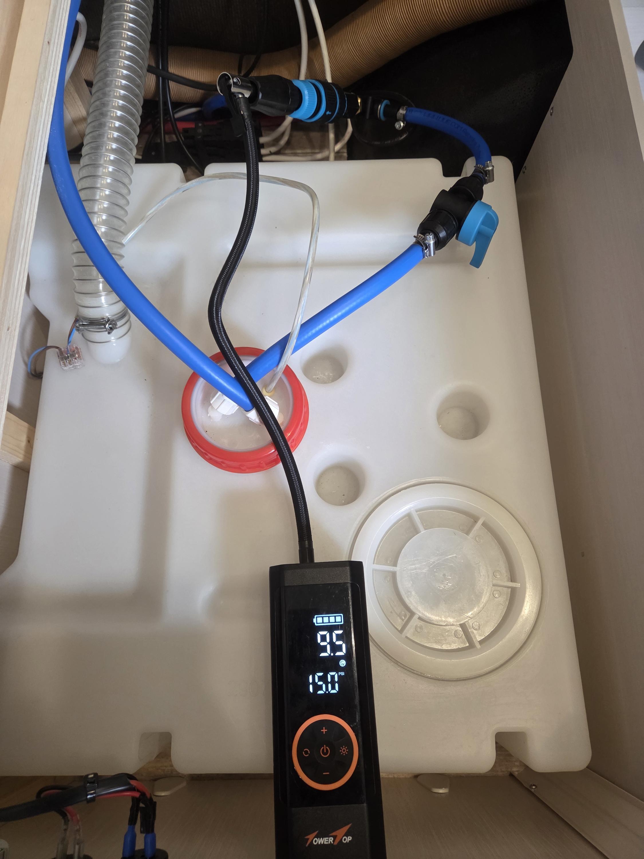Wei&szlig;e Kunststoffwanne mit runden Vertiefungen, mehrere Schl&auml;uche und Kabel (blau, schwarz, silber) oben, schwarzes Handger&auml;t mit beleuchtetem Display und orangefarbener Umrandung in der Wanne, roter Dichtungsring neben einem Schlauch, runder Ablaufde