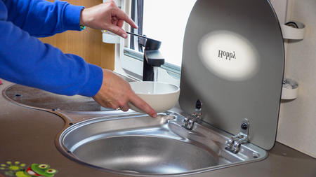 Zwei H&auml;nde &uuml;ber einem runden Edelstahlsink, eine Hand h&auml;lt eine wei&szlig;e Sch&uuml;ssel unter einem Wasserhahn, die andere zeigt auf den Hahn; ge&ouml;ffneter grauer Klappdeckel mit Aufschrift "HiPP", Fenster im Hintergrund
