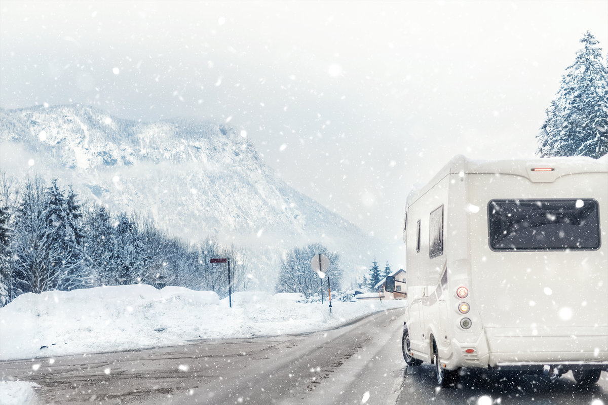 Heckansicht eines wei&szlig;en Wohnmobils auf einer schneebedeckten Stra&szlig;e, fallender Schnee, verschneite Tannen und Berge im Hintergrund