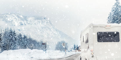 Heckansicht eines wei&szlig;en Wohnmobils auf einer schneebedeckten Stra&szlig;e, fallender Schnee, verschneite Tannen und Berge im Hintergrund