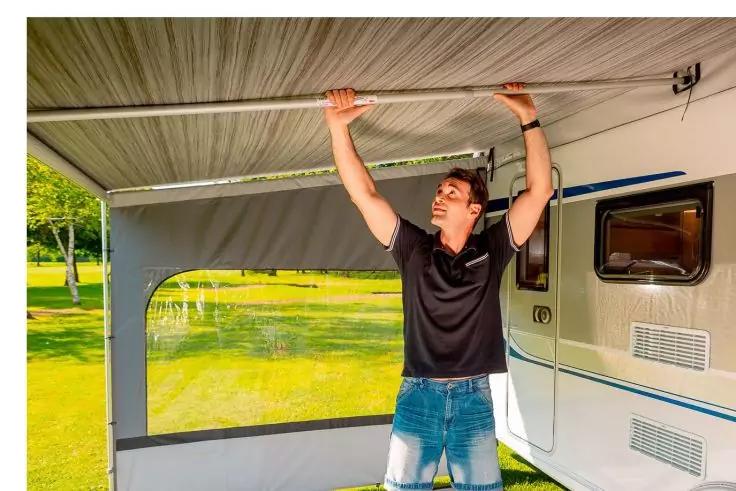 Homme en polo noir et jean tenant une barre horizontale au-dessus de sa tête sous un auvent à côté d'une caravane ; pelouse et arbres en arrière-plan