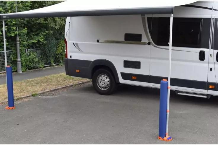 Camping-car blanc de côté, store déployé avec deux poteaux de soutien bleus sur ancrages au sol orange, surface de stationnement goudronnée et haie en arrière-plan