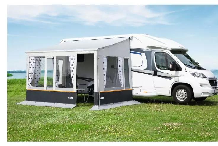 Camping-car blanc avec un auvent gris fixé sur un pré, eau et ciel en arrière-plan