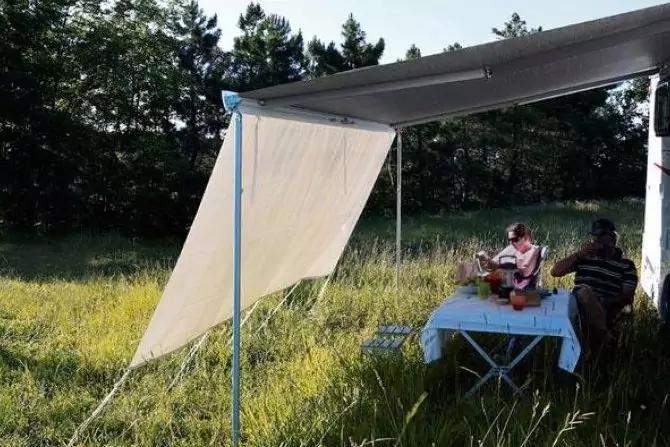 Deux personnes assises à une table avec une nappe blanche sous une tonnelle dans un champ herbeux ; une personne tient une guitare ; arbres en arrière-plan