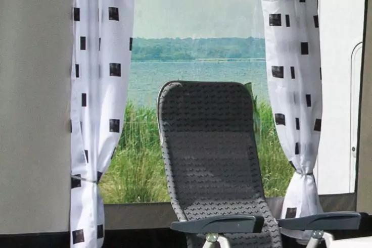 Ouverture de fenêtre avec rideaux blancs à carrés noirs ; devant un fauteuil-long en osier gris, herbe, un lac et une ligne d'arbres au loin