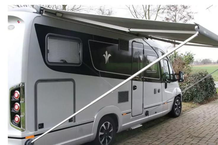 Camping-car blanc et gris avec auvent déployé sur un emplacement pavé, arbres et champ en arrière-plan