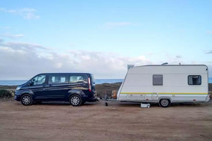 Fourgon noir avec caravane blanche attel&eacute;e sur un terrain non goudronn&eacute; au bord de la mer ; c&ocirc;te et ciel nuageux en arri&egrave;re-plan