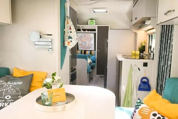 Intérieur avec table ronde, plateau avec verre et plante, coussins colorés sur banquettes, coin cuisine avec évier, citrons et placards, couloir vers coin nuit avec carte au mur