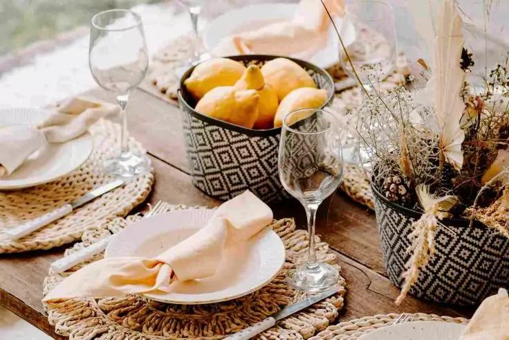 Table en bois avec sets tissés, assiettes blanches, serviettes en tissu claires, verres à vin transparents, bol à motif avec poires jaunes et pots à motif contenant herbes séchées
