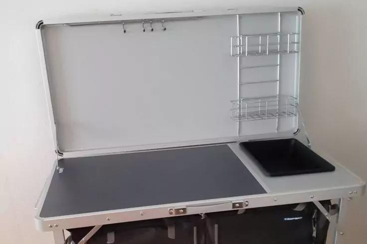 Mallette pliable argentée ouverte avec grande surface grise à gauche et petit bac noir encastré à droite ; intérieur du couvercle avec trois crochets en haut et étagères en fil métallique à droite ; pieds pliants et mur blanc en arrière-plan