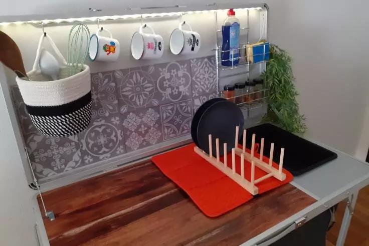 Plan de travail en bois, rail métallique avec quatre tasses suspendues, panier suspendu tressé blanc avec ustensiles en bois, crédence grise à motifs, tapis rouge et noir, support en bois avec chevilles verticales, plateau noir, bouteille bleue sur petite étagère, plante verte suspendue