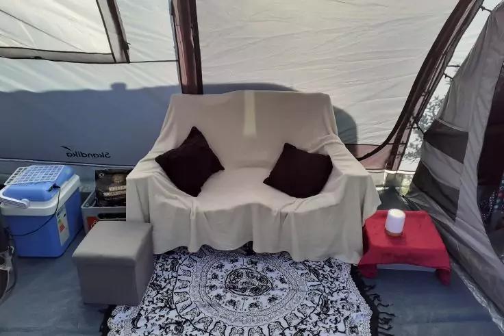 Banquette deux places recouverte d'un drap blanc avec deux coussins foncés, tapis noir et blanc à motifs devant, glacière bleue et pouf gris à gauche, petite table d'appoint rouge avec une tasse blanche à droite, parois textiles claires et courbées avec fenêtres transparentes en arrière-plan