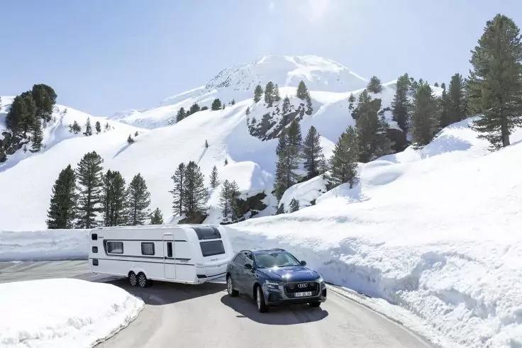 Voiture avec caravane attelée sur une route de montagne enneigée, hautes congères, conifères et sommet enneigé en arrière-plan