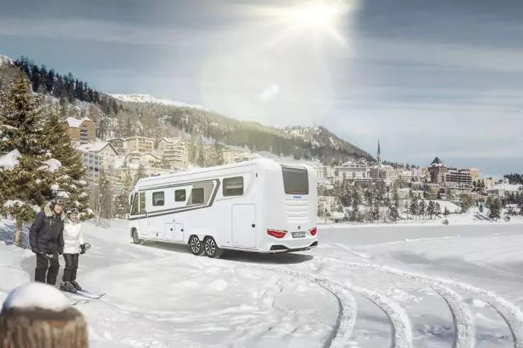 Camping-car sur une &eacute;tendue enneig&eacute;e&nbsp;; deux personnes en tenue d'hiver pr&egrave;s d'un poteau en bois, maisons enneig&eacute;es sur la colline, soleil dans le ciel, traces de pneus dans la neige