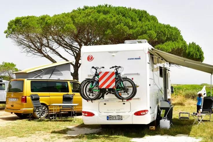 Vue arri&egrave;re d'un camping-car blanc avec deux v&eacute;los sur un porte-v&eacute;los et un panneau rouge et blanc, auvent d&eacute;ploy&eacute; ; &agrave; gauche un van jaune, pins en arri&egrave;re-plan, chaises pliantes et une personne debout &agrave; droite