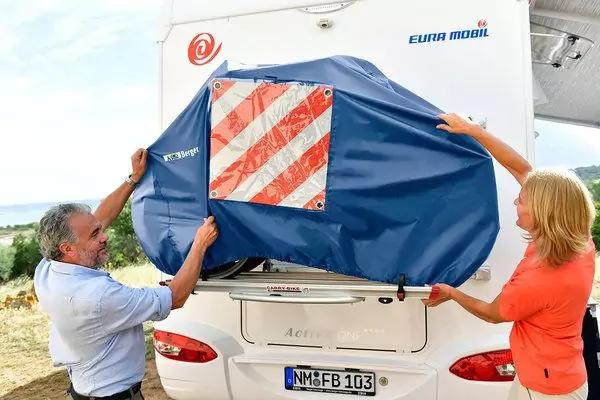 Deux personnes tirent une housse bleue sur l'arri&egrave;re d'une caravane blanche&nbsp;; la housse comporte un panneau d'avertissement &agrave; bandes diagonales rouge et blanc
