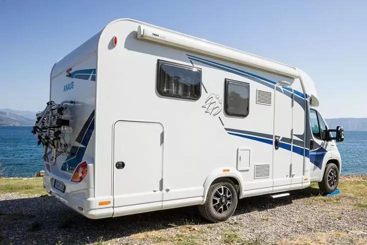 Camping-car blanc avec bandes bleues et vélos sur un porte-vélos arrière, garé sur du gravier au bord de la mer, montagnes en arrière-plan