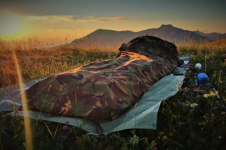 Personne dans un sac de couchage camouflage sur un matelas de sol dans une prairie fleurie, montagnes en arrière-plan, soleil juste au-dessus de l'horizon