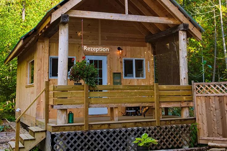 Petite cabane en bois avec véranda dans une forêt, panneau "Rezeption" au-dessus de la porte, deux fenêtres, plante suspendue, arrosoir sur la véranda