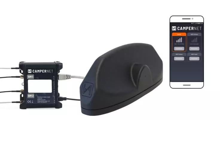 Appareil noir allongé et arrondi sur fond blanc, à sa gauche un boîtier rectangulaire noir avec des câbles, à sa droite un smartphone affichant un écran d'application avec 'CAMPERNET', des barres de signal et des boutons visibles