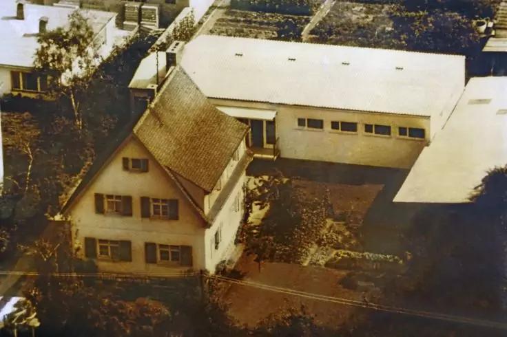 Vue a&eacute;rienne de deux b&acirc;timents c&ocirc;te &agrave; c&ocirc;te : &agrave; gauche une maison blanche &agrave; plusieurs &eacute;tages avec toit en pente et volets, &agrave; droite un b&acirc;timent long &agrave; toit plat avec rang&eacute;es de fen&ecirc;tres ; devant une cour pav&eacute;e avec buissons et arbres. Photo en ton s&eacute;pia