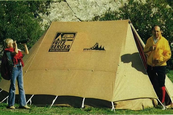 Tente beige avec logo FRITZ BERGER imprim&eacute; sur une pelouse; une personne &agrave; gauche et une personne &agrave; droite; arbres en arri&egrave;re-plan