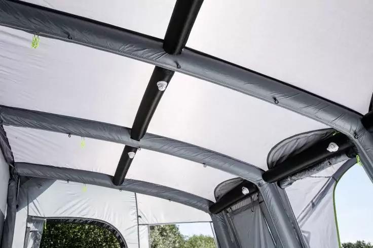 Vue intérieure avec plafond en tissu blanc incurvé, plusieurs profilés tubulaires gris courbés, parois latérales en tissu avec ouvertures de fenêtres et arbres verts visibles par une ouverture