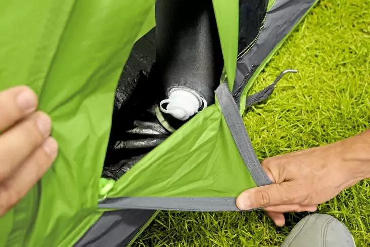 Deux mains tiennent ouverte une ouverture de tente verte, à l'intérieur un objet en plastique blanc sur un sol intérieur noir, herbe verte à côté