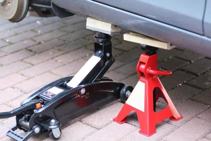 Cric nero e cavalletto rosso con blocchetti di legno sotto un'auto su pavimentazione in pietra