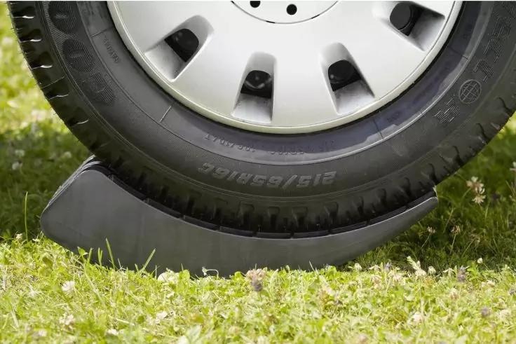 Primo piano di una gomma d'auto con copriruota argentato accanto a un cuneo nero per ruota su erba con piccoli fiori bianchi