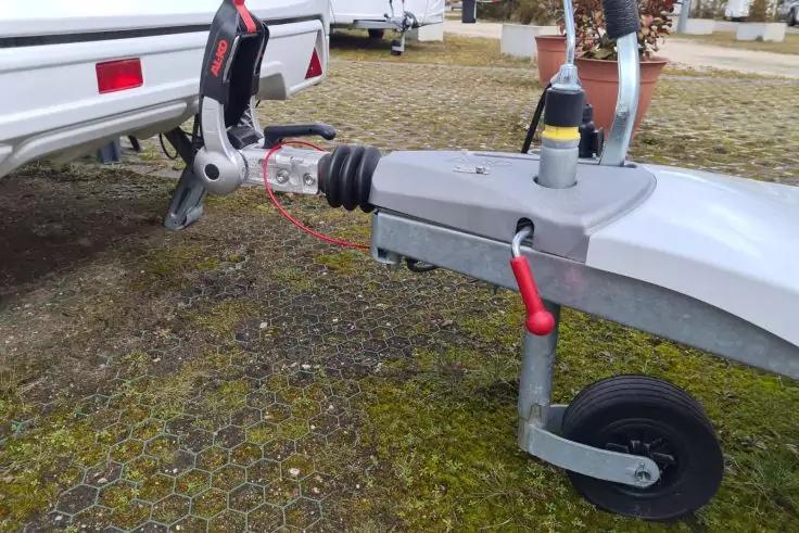 Primo piano del gancio di traino di una roulotte con ruota di supporto, testa di accoppiamento, maniglia rossa e cavo di sicurezza su pavimentazione muschiosa