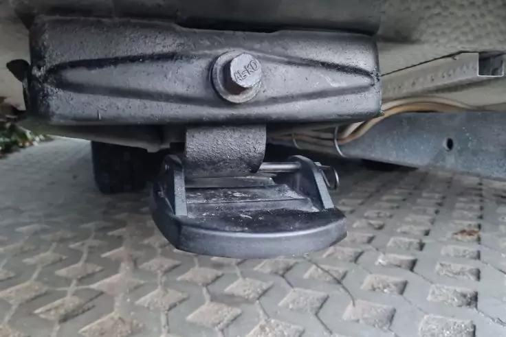 Supporto metallico nero con vite esagonale visibile e tampone rotondo in gomma nera sotto un veicolo, rivestimento grigio sopra; pavimentazione acciottolata