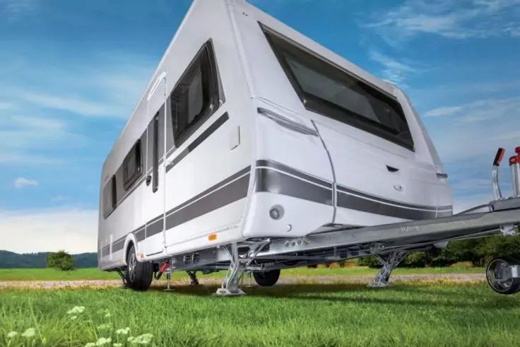 Caravan bianco su erba verde, piedi di supporto anteriori e barra di traino visibili, finestre, cielo azzurro con nuvole, colline verdi sullo sfondo