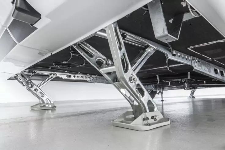 Vista inferiore di una piattaforma con pi&ugrave; supporti metallici argentati incrociati fissati a una superficie inferiore nera e liscia, su un pavimento chiaro