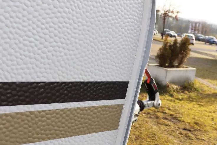 Primo piano di un pannello bianco strutturato con strisce orizzontali nere e beige; bordo metallico a destra, sullo sfondo erba, un vaso con arbusti e auto parcheggiate