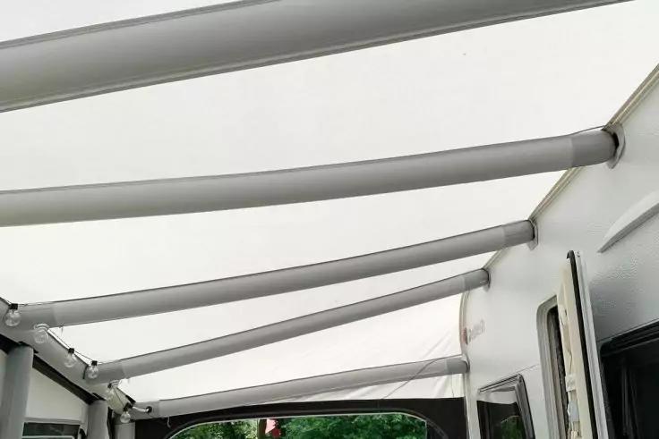 Tendalino in tessuto bianco con travi di sostegno orizzontali fissato a un veicolo con porta e finestra, alberi sullo sfondo