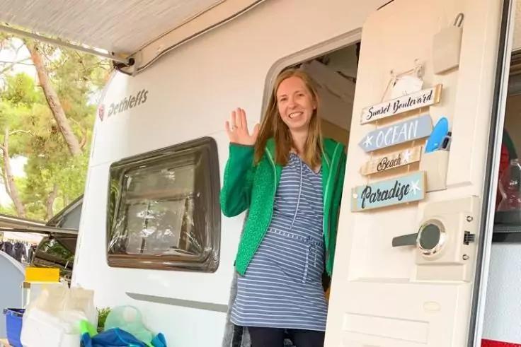 Persona in piedi nella porta aperta di un caravan, saluta con la mano, indossa un cardigan verde e un vestito a righe; tendalino aperto; borse e contenitori accanto all'ingresso; cartelli decorativi in legno sulla porta