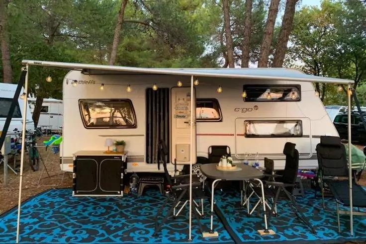 Roulotte bianca con tenda estesa; in primo piano telo da terra blu a motivo, tavolo pieghevole con sedie, mobile contenitore scuro, tazze e bottiglie sul tavolo, alberi sullo sfondo