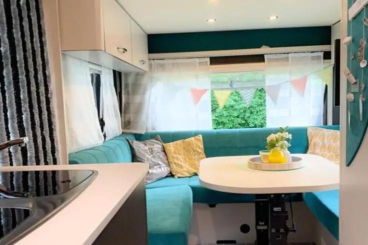 Divano a L turchese con cuscini gialli, tavolo rotondo bianco con vaso di fiori gialli e un limone, finestra con tende bianche e bandierine colorate, piano cucina con lavello a sinistra, pensili bianchi