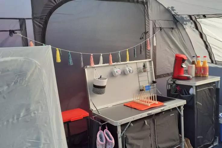 Interno di una tenda con tavolo pieghevole, scolapiatti rosso, tazze appese, corda con nappine colorate, sgabello rosso, macchina da caff&egrave; rossa, tre bottiglie di succo e pantofole rosa