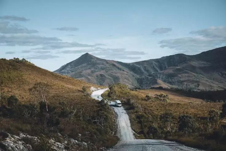 Strada sterrata che serpeggia attraverso paesaggio collinare con bassa vegetazione verso le montagne; un veicolo sulla strada; cielo nuvoloso