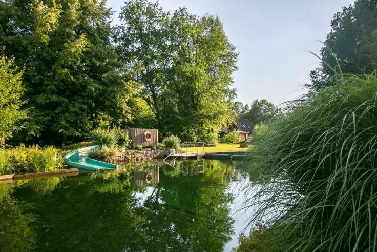 Laghetto con scivolo verde nell'acqua, pontile in legno, salvagente, erba sulla riva, alberi e riflesso
