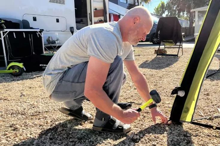 Persona inginocchiata con maglietta grigia, che usa un martello con manico giallo-nero per conficcare un picchetto metallico in terreno ghiaioso accanto a un oggetto nero e verde; camper e attrezzatura sullo sfondo