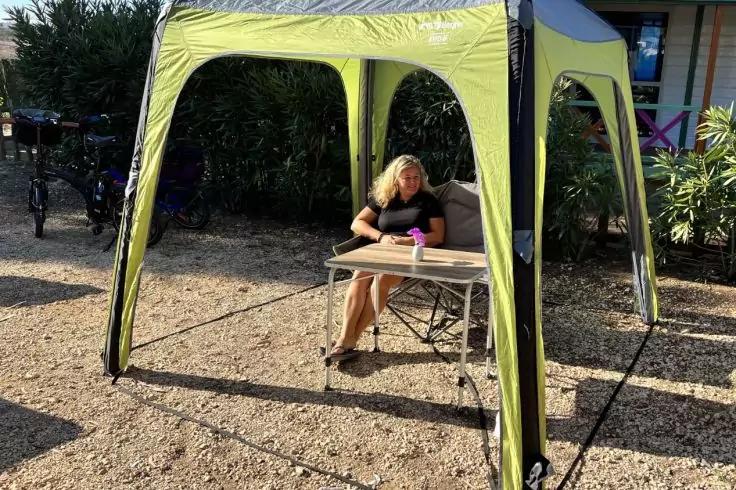 Persona seduta a un tavolo pieghevole sotto un padiglione verde; tazza rosa sul tavolo; terreno ghiaioso, cespugli e biciclette sullo sfondo